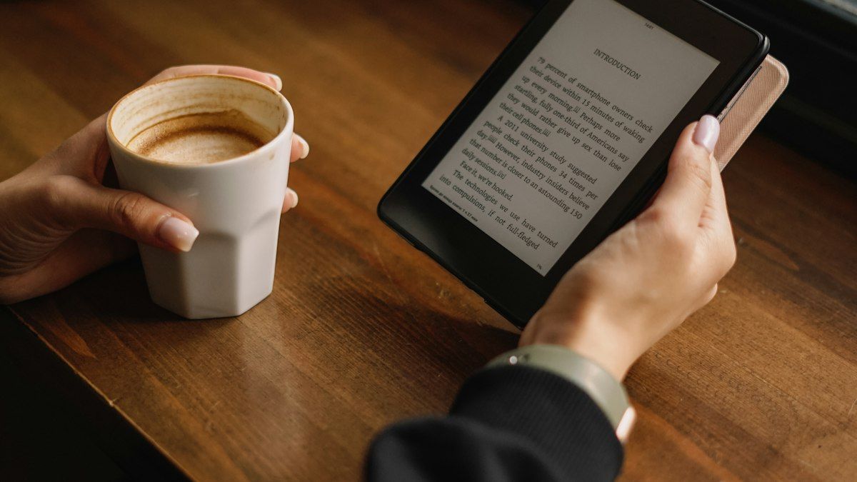 Tangan memegang e-reader di samping secangkir kopi