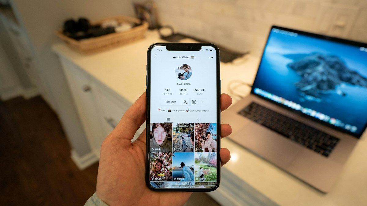 Tangan memegang smartphone dengan profil TikTok di depan laptop