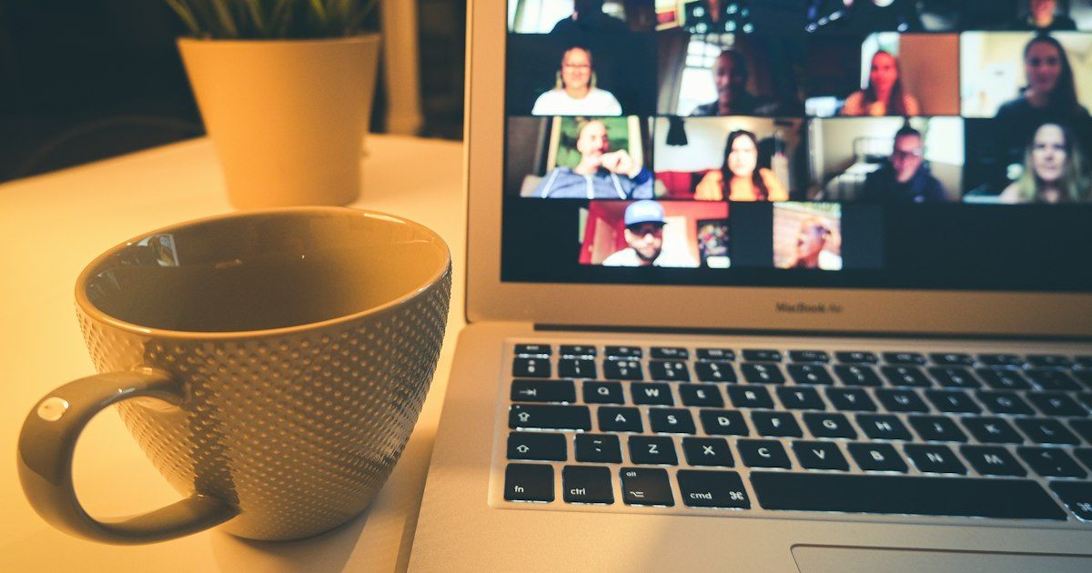 Laptop menampilkan rapat video dengan banyak peserta di samping cangkir kopi